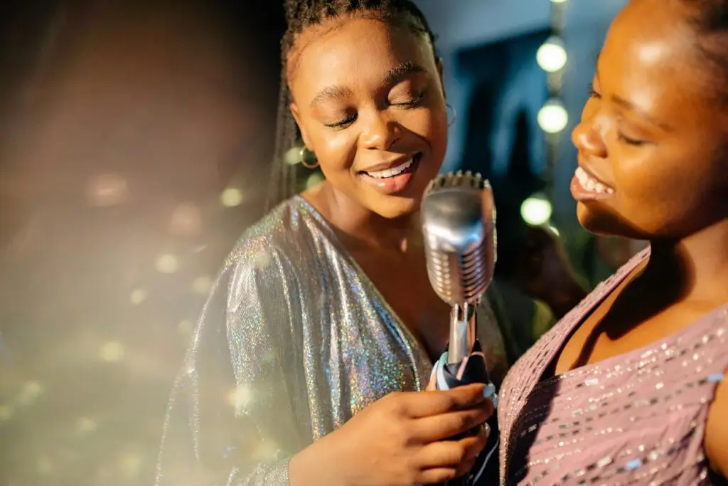 Two women joyfully singing together with a vintage microphone in a lively, indoor setting.